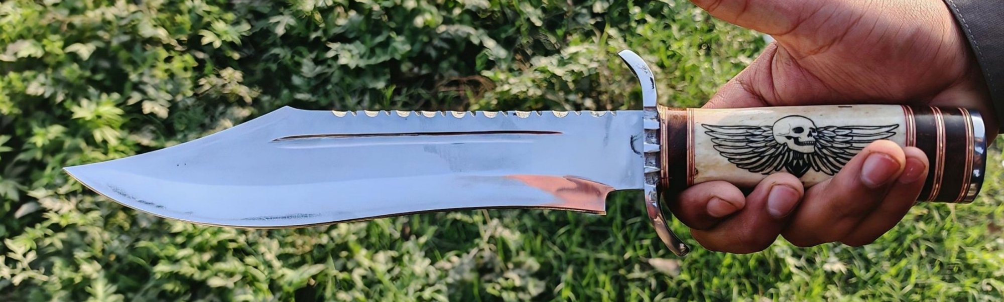 Bowie knife with a long clip point blade, serrated spine and bone handle engraved with a winged skull