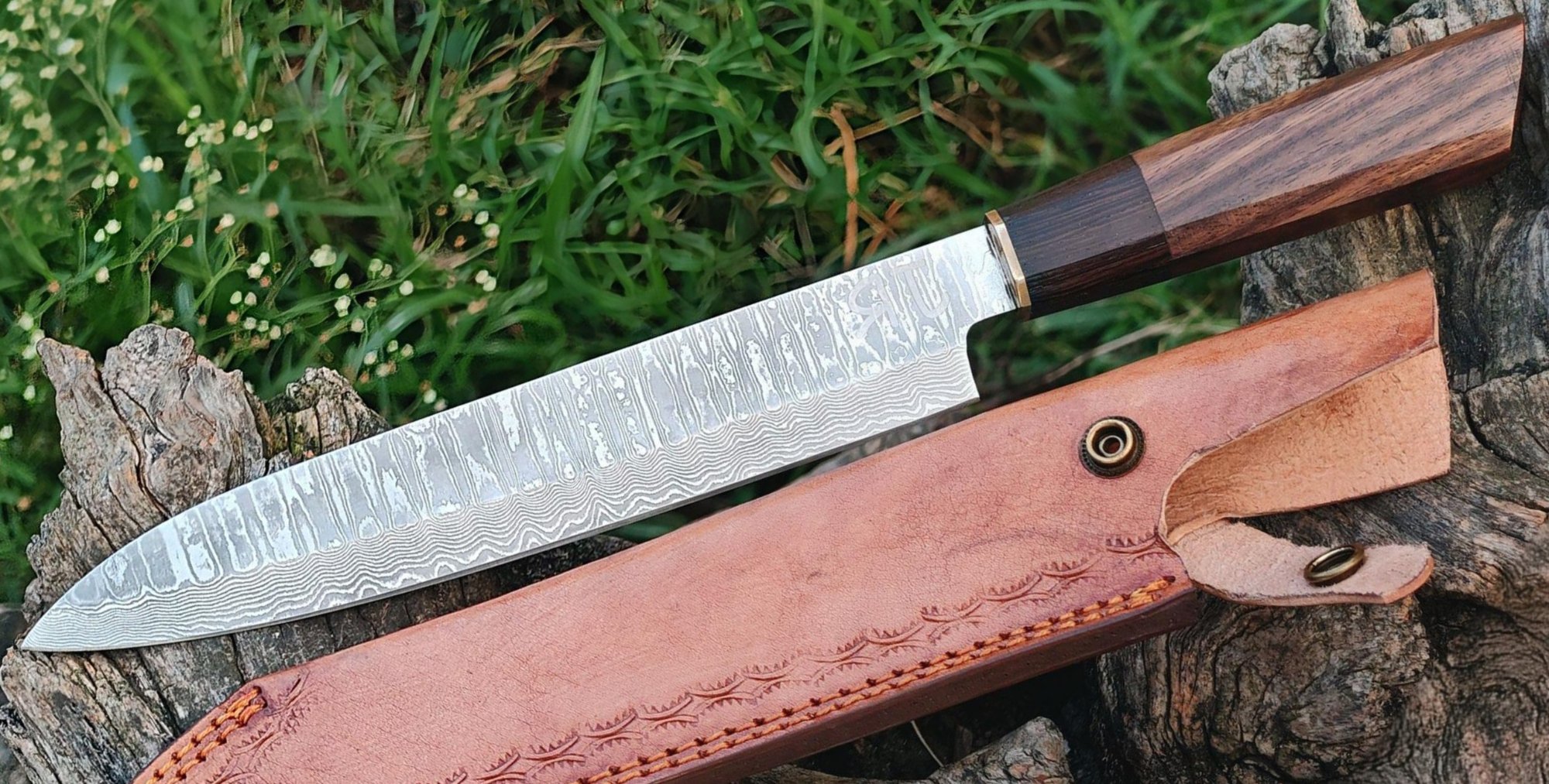 Damascus chef knife with a rosewood handle beside a tooled leather sheath on a tree stump