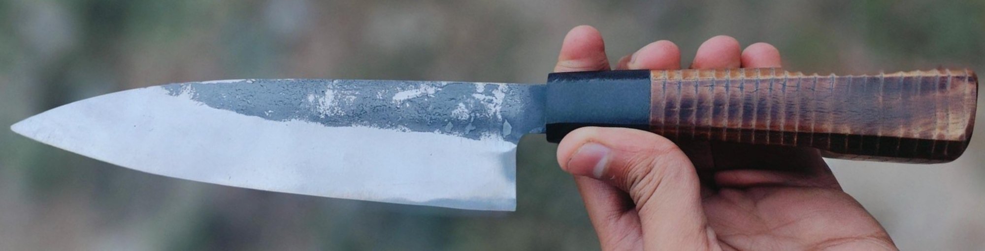 Kiritsuke chef knife with a dark hammered finish and a scorched wood handle, held in hand